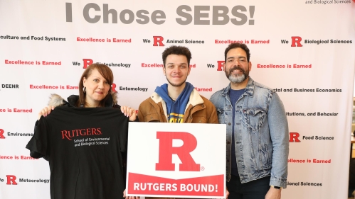 An incoming SEBS student and two parents, standing under a sign that says, 'I Choose SEBS!'.